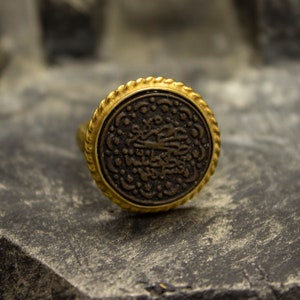 May include: A gold ring with a black circular design featuring intricate Arabic calligraphy. The ring has a rope-like border.