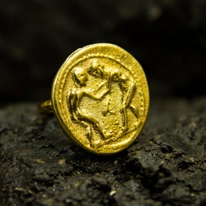 May include: A gold ring with a detailed design of two figures holding hands. The ring is set against a dark background.
