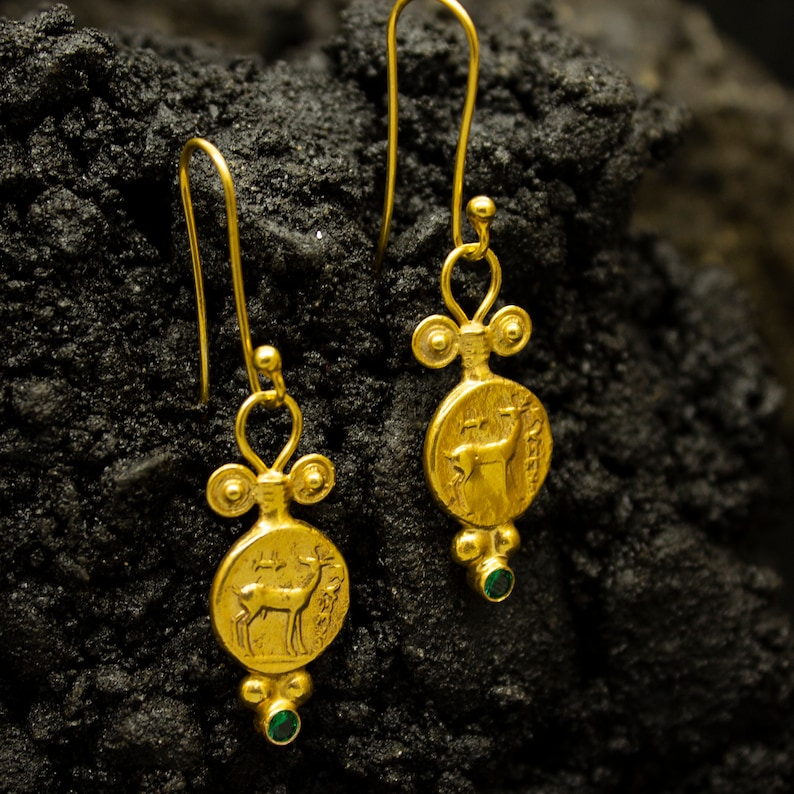 Pu&ograve; includere: Un paio di orecchini pendenti in oro con un accento di pietra preziosa verde. Ogni orecchino presenta un ciondolo rotondo con un'immagine dettagliata di un cervo.