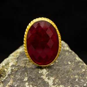 Può includere: Un anello dorato con una grande gemma rosso scuro, sfaccettata e di forma ovale. La gemma è incastonata in una fascia dorata testurizzata a forma di corda. L'anello è esposto su una superficie grigia strutturata su sfondo nero.