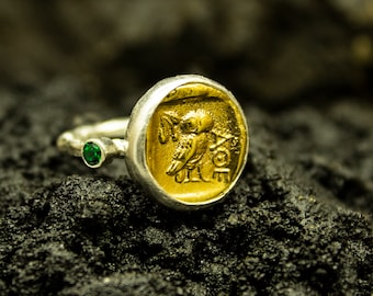 May include: A silver ring with a gold owl design and a green gemstone accent. The ring is set against a dark, textured background.