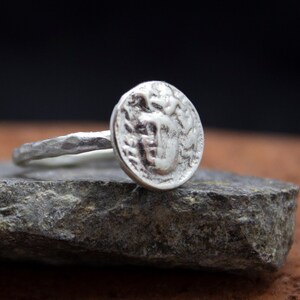 Op de afbeelding: Een zilveren ring met een getextureerde band en een ronde, muntvormige bovenkant met een reli&euml;fdesign.