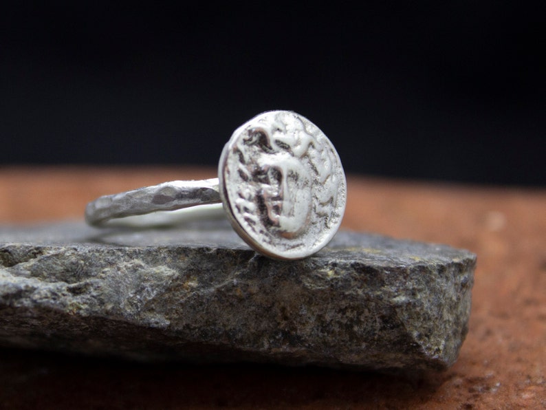 Op de afbeelding: Een zilveren ring met een getextureerde band en een ronde, muntvormige bovenkant met een reli&euml;fdesign.