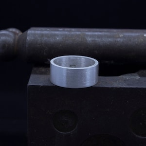 Puede incluir: Un anillo de plata con acabado cepillado. El anillo es grueso y tiene una parte superior plana. El anillo está marcado con el número "925" en el interior.