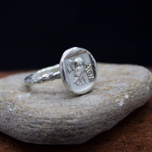 May include: A silver ring with a textured band and a round, hammered silver top featuring a detailed image of a bird and the inscription "AOE".