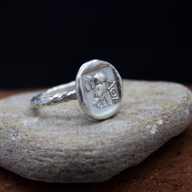 May include: A silver ring with a textured band and a round, hammered silver top featuring a detailed image of a bird and the inscription "AOE".