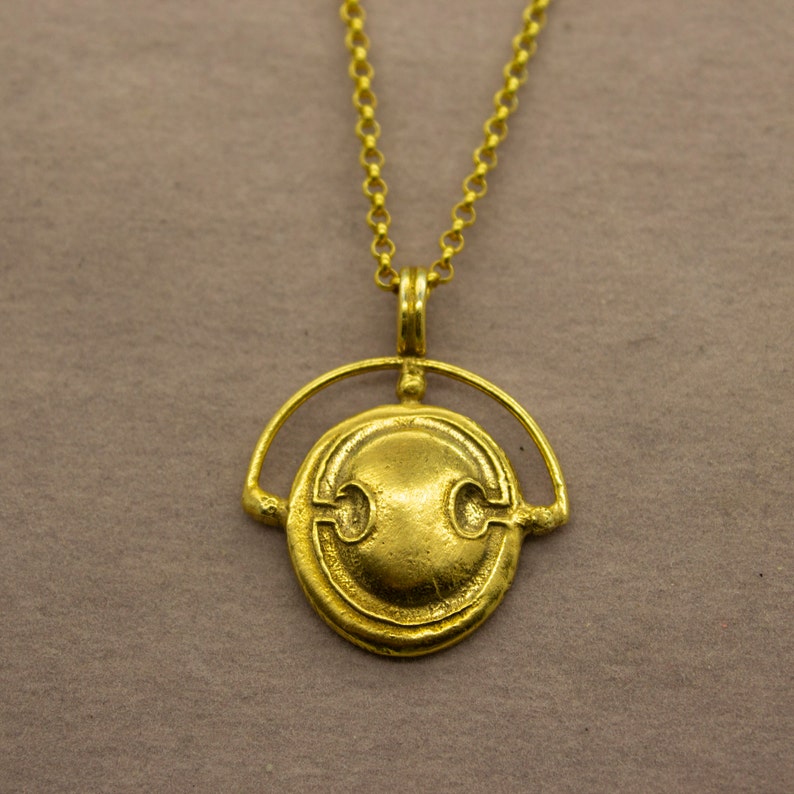 Puede incluir: Un collar de oro con un colgante redondo que presenta un dise&ntilde;o estilizado. El colgante est&aacute; suspendido de una cadena delicada.