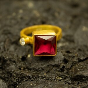 Peut inclure: Une bague en or ornée d'une grande pierre rouge rubis carrée. Une petite pierre ronde et transparente est sertie sur le côté. La bague a une finition mate et texturée.
