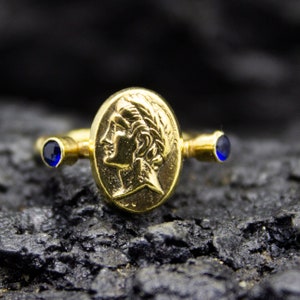 May include: A gold ring with a blue gemstone on each side and a raised image of a woman's face in the center.
