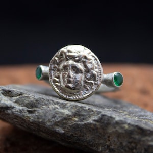 May include: A silver ring with a round, detailed design featuring a face with a crown of snakes. The ring has two green gemstones on either side of the design.