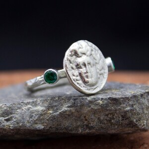 May include: A silver ring with a textured band and a round coin-shaped design featuring a raised image. The ring is accented with two green gemstones.