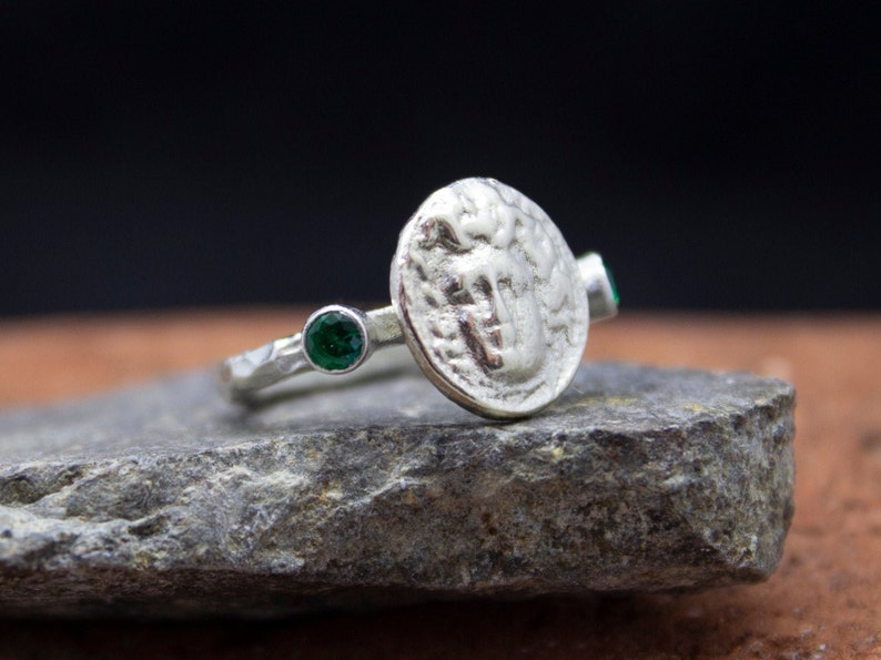 May include: A silver ring with a textured band and a round coin-shaped design featuring a raised image. The ring is accented with two green gemstones.