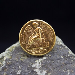 Peut inclure: Une pi&egrave;ce de monnaie dor&eacute;e avec une image en relief d'une figure ail&eacute;e assise sur un tr&ocirc;ne. La pi&egrave;ce a un design d&eacute;taill&eacute; et une surface lisse et brillante.