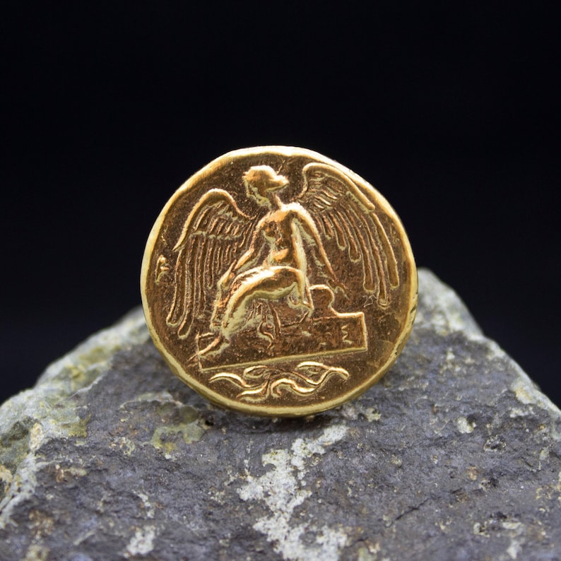 Peut inclure: Une pi&egrave;ce de monnaie dor&eacute;e avec une image en relief d'une figure ail&eacute;e assise sur un tr&ocirc;ne. La pi&egrave;ce a un design d&eacute;taill&eacute; et une surface lisse et brillante.