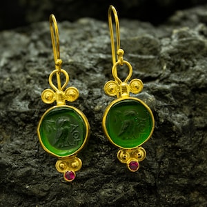 May include: A pair of gold earrings with green glass insets featuring an engraved bird design. Each earring has a small ruby-colored stone at the bottom.