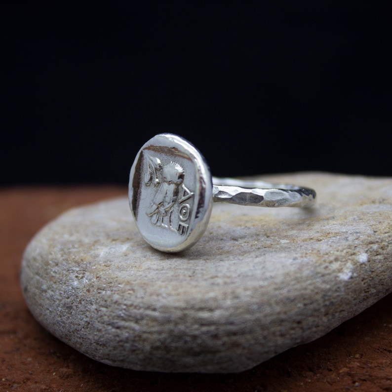 May include: A silver ring with a textured band and a round, hammered silver top featuring a raised design of a winged figure holding a staff.