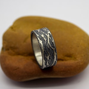 May include: A silver ring with a black oxidized finish. The ring has a textured design that resembles tree bark.