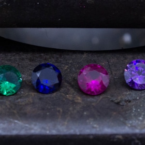 Pu&ograve; includere: Sette pietre preziose rotonde di vari colori, tra cui bianco, verde, blu e viola, sono disposte in fila su una superficie grigio scuro.