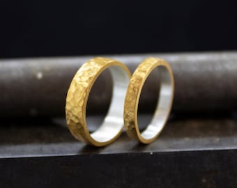 May include: Two gold wedding bands with a hammered texture. The bands are sitting on a dark gray surface.
