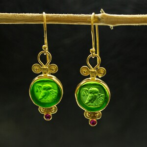 May include: Gold earrings with green glass owl charms and pink gemstones. The owl charms are detailed with a wing and a branch.
