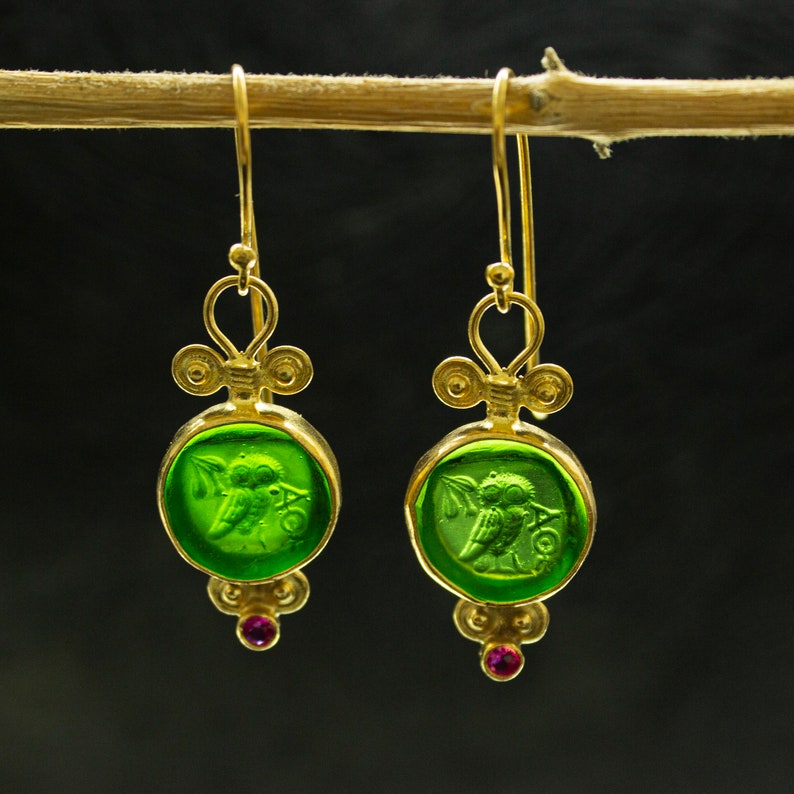 May include: Gold earrings with green glass owl charms and pink gemstones. The owl charms are detailed with a wing and a branch.