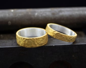 May include: Two gold wedding bands with a hammered texture. The bands are stacked on top of each other, with the top band slightly offset to the right. The bands are on a dark gray surface.