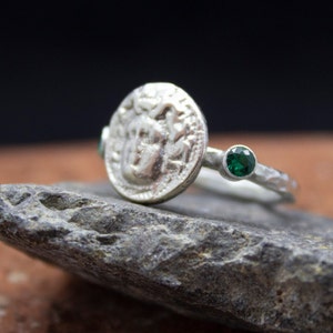 May include: A silver ring with a textured band and a round coin-shaped design featuring a face. Two green gemstones are set on either side of the coin.