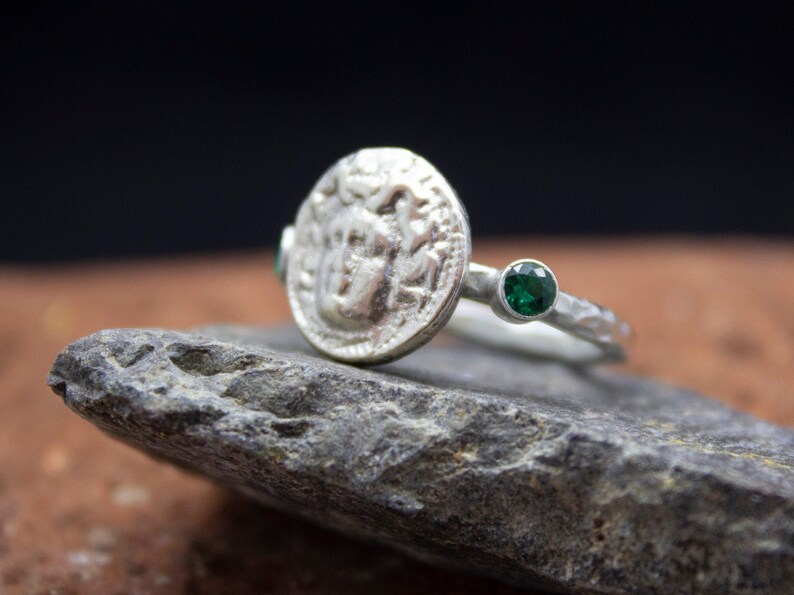 May include: A silver ring with a textured band and a round coin-shaped design featuring a face. Two green gemstones are set on either side of the coin.
