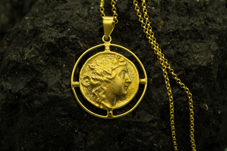 May include: A gold necklace with a round pendant featuring a profile of a woman's face. The pendant is surrounded by a decorative circle.