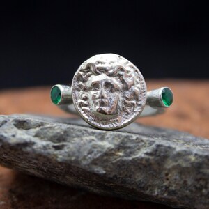May include: A silver ring with a round, detailed design of a face with a crown of snakes. The ring has two green gemstones on either side of the face.
