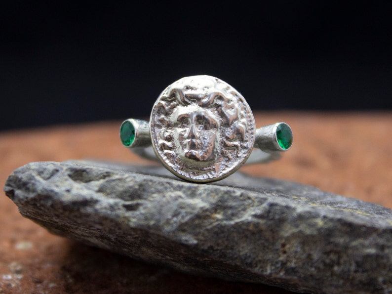 May include: A silver ring with a round, detailed design of a face with a crown of snakes. The ring has two green gemstones on either side of the face.