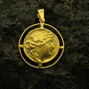 May include: A gold pendant featuring a detailed profile of a woman's face with a laurel wreath. The pendant is framed by a circular design with four open spaces.