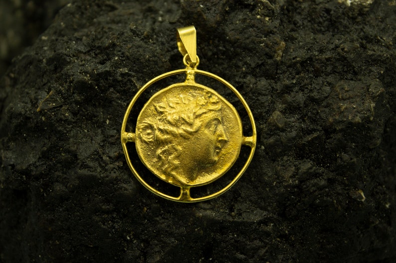 May include: A gold pendant featuring a detailed profile of a woman's face with a laurel wreath. The pendant is framed by a circular design with four open spaces.