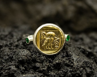 May include: A silver ring with a gold coin-shaped setting featuring an owl design. The ring has two green gemstones on either side of the setting.