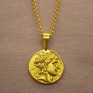 May include: A gold chain necklace with a pendant featuring a detailed profile of a person's head. The pendant is oval-shaped and has a textured surface.