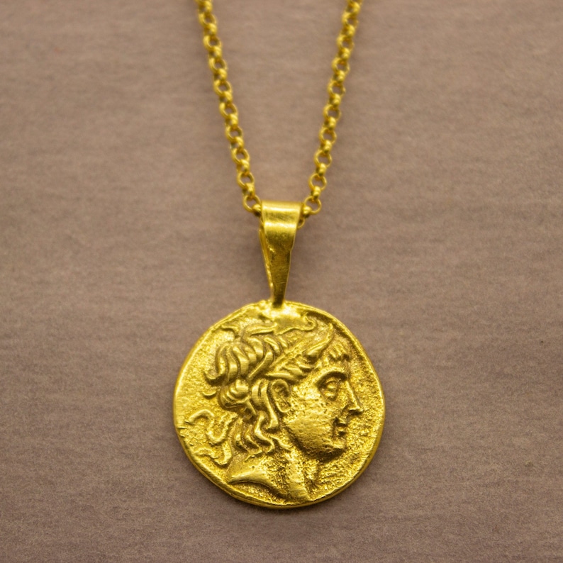 May include: A gold chain necklace with a pendant featuring a detailed profile of a person's head. The pendant is oval-shaped and has a textured surface.