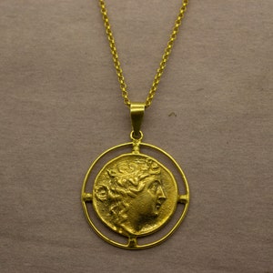 May include: A gold necklace with a round pendant featuring a profile of a woman's face with a laurel wreath. The pendant is surrounded by a circular frame with four decorative accents.