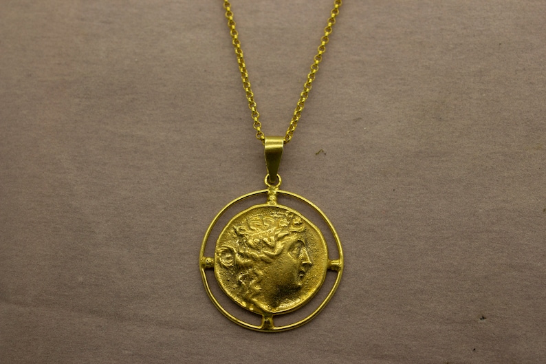 May include: A gold necklace with a round pendant featuring a profile of a woman's face with a laurel wreath. The pendant is surrounded by a circular frame with four decorative accents.