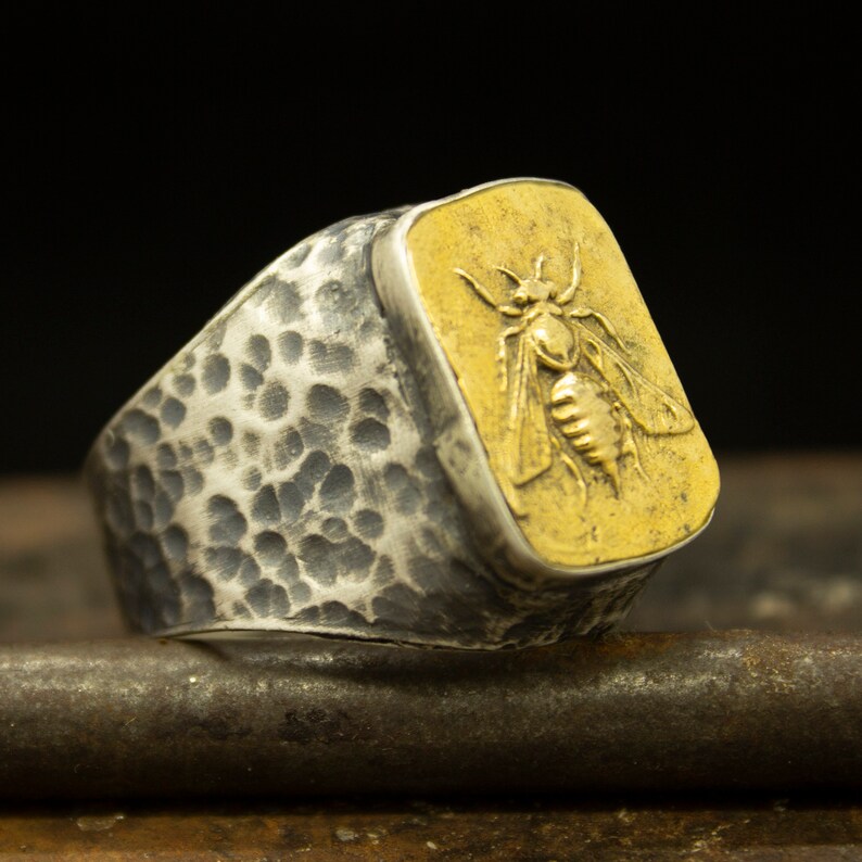 May include: A silver ring with a hammered texture and a gold square inset featuring a detailed bee design.