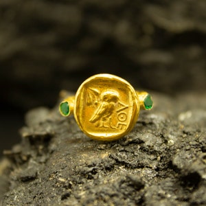 May include: A gold ring with a green stone on each side. The center of the ring features a raised design of a bird with the word "AOE" inscribed below it.