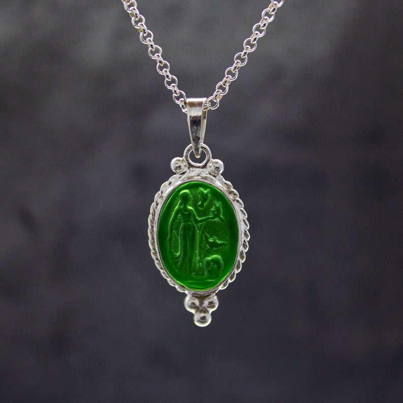 Puede incluir: Un collar de cadena de plata con un colgante ovalado verde que presenta un dise&ntilde;o en relieve de dos figuras. El colgante tiene un borde de plata detallado.