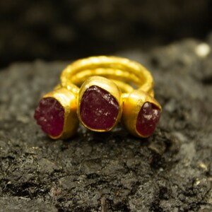 Puede incluir: Un anillo de oro con tres piedras de rubí rojo engastadas en la banda. El anillo está sobre una superficie oscura y texturizada.