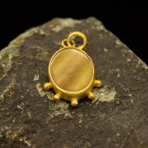 May include: A gold-colored pendant with a circular, light brown stone center. The pendant has a loop at the top for a chain and four decorative, rounded protrusions. The pendant is displayed on a dark, textured surface.
