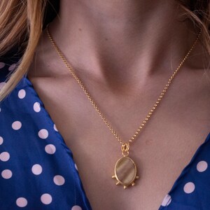 May include: A gold-colored necklace with a pendant featuring an oval-shaped, light-colored stone. The pendant is adorned with small, round embellishments. The necklace is worn over a blue dress with white polka dots.