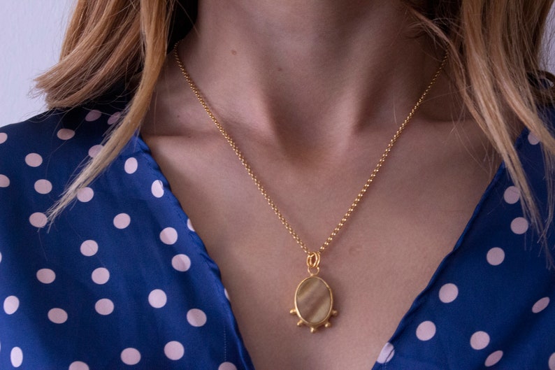 May include: A gold-colored necklace with a pendant featuring an oval-shaped, light-colored stone. The pendant is adorned with small, round embellishments. The necklace is worn over a blue dress with white polka dots.