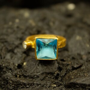 Könnte beinhalten: Ein goldener Ring mit einem quadratischen blauen Edelstein. Der Ring liegt auf einer dunklen, strukturierten Oberfläche.