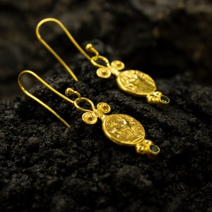 Pu&ograve; includere: Un paio di orecchini d'oro con un design a forma di moneta e pietre preziose verdi. Le monete presentano un design in rilievo di un cavallo e un cavaliere.