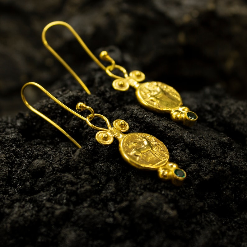 Pu&ograve; includere: Un paio di orecchini d'oro con un design a forma di moneta e pietre preziose verdi. Le monete presentano un design in rilievo di un cavallo e un cavaliere.