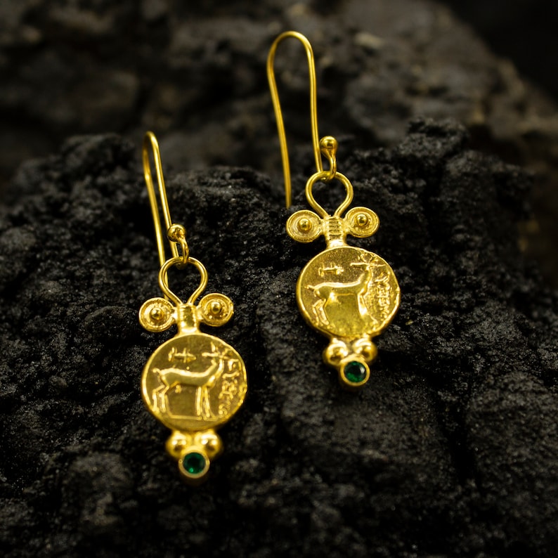 Pu&ograve; includere: Orecchini pendenti in oro con un ciondolo a forma di moneta che presenta un design a cervo e accenti di pietre preziose verdi.
