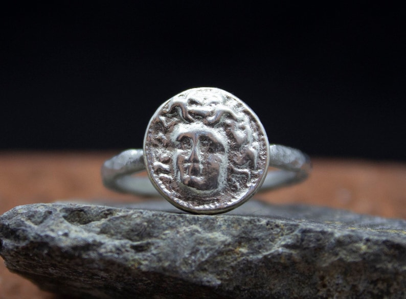 Op de afbeelding: Een zilveren ring met een ronde, getextureerde band en een groot, rond, muntvormig ontwerp met een gedetailleerd profiel van Medusa met slangen in haar haar.